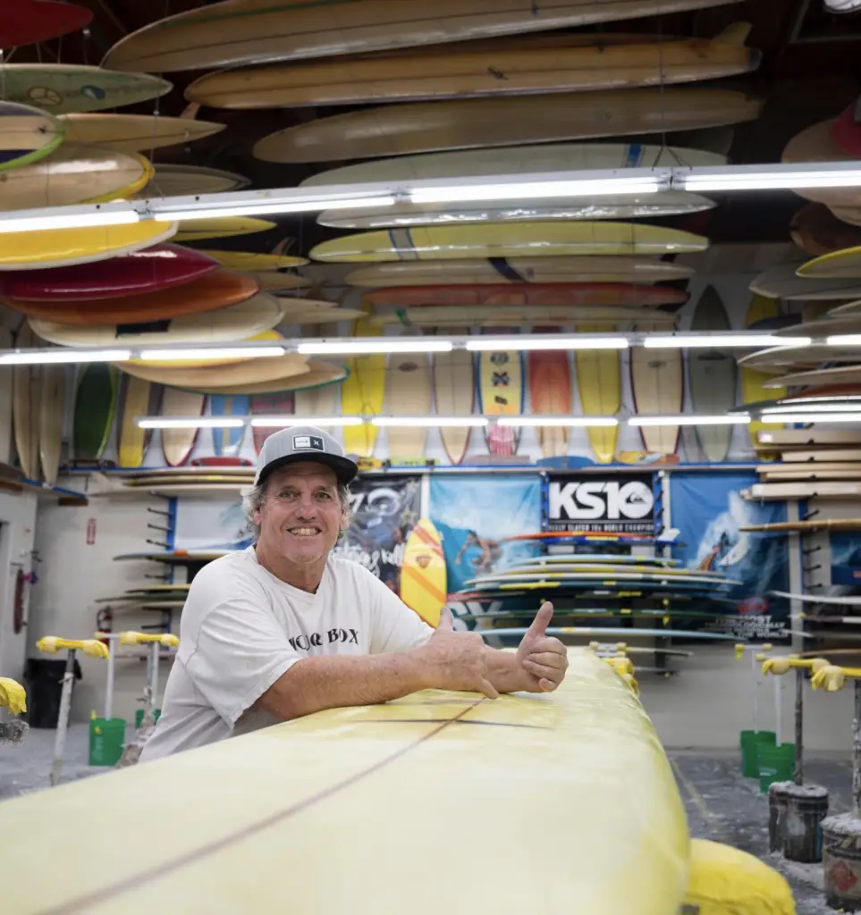 Joe Roper at his surfboard factory smiling with a yellow surfboard and his vintage surfboard collection in the background.