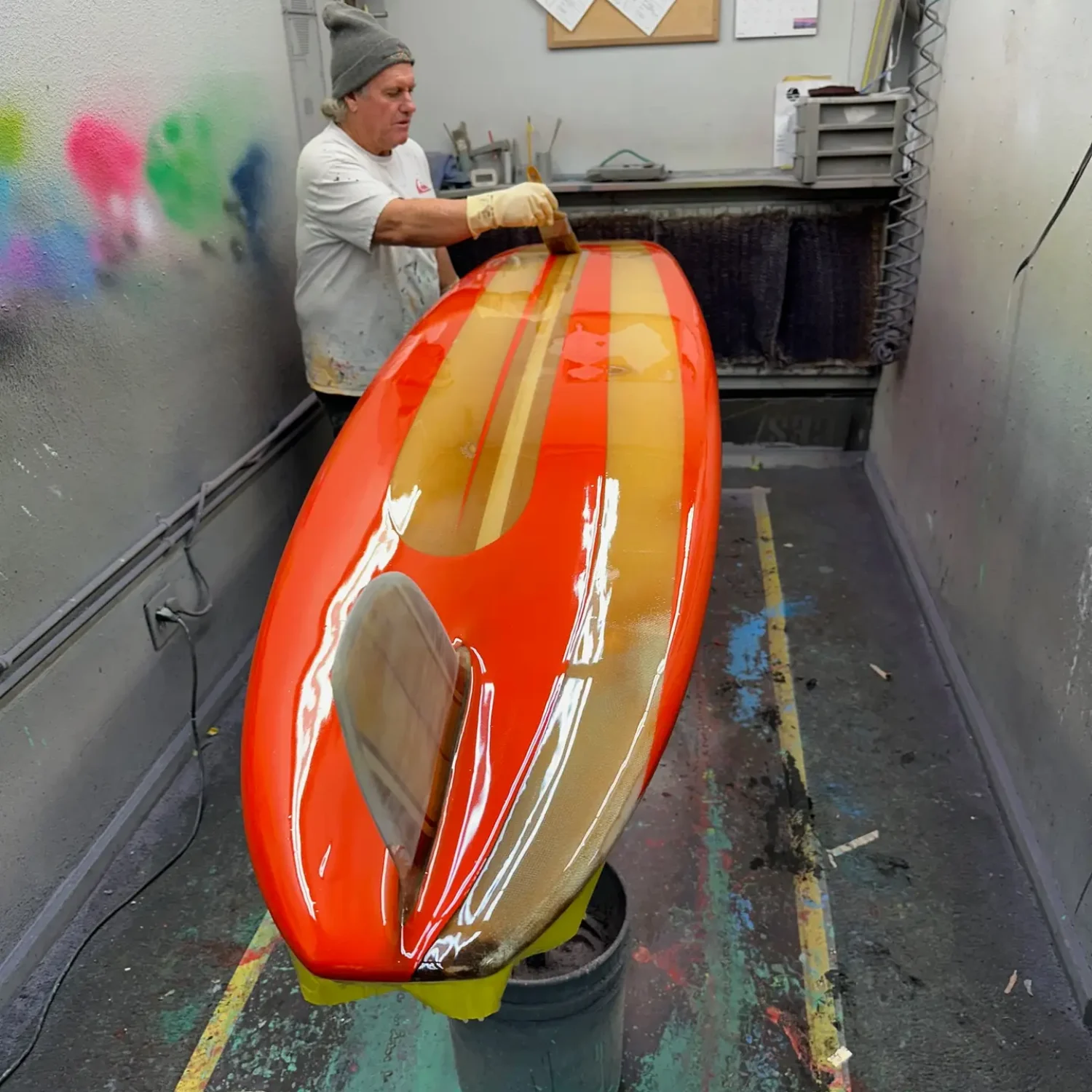 Joe Roper working in his factory on a vintage longboard surfboard that is red and wood with a big glass on wooden fin.